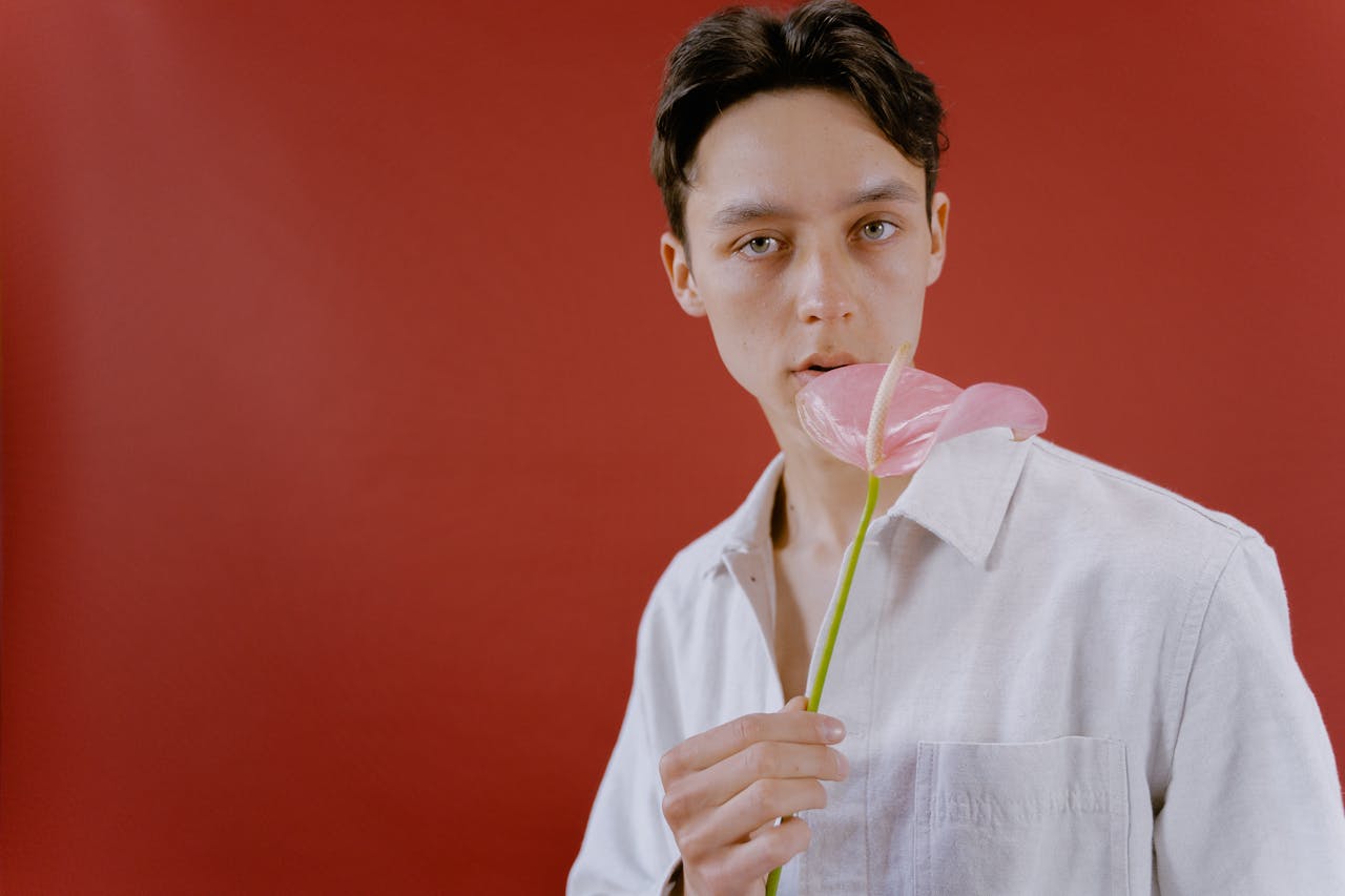 A young adult in a white shirt holding a pink anthurium against a red backdrop.